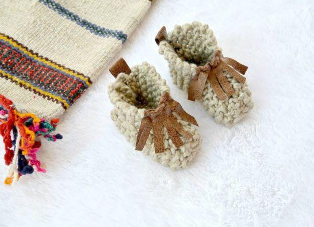 Cream knitted baby booties with suede fringe and leather ties, on a white fluffy rug beside a striped cushion