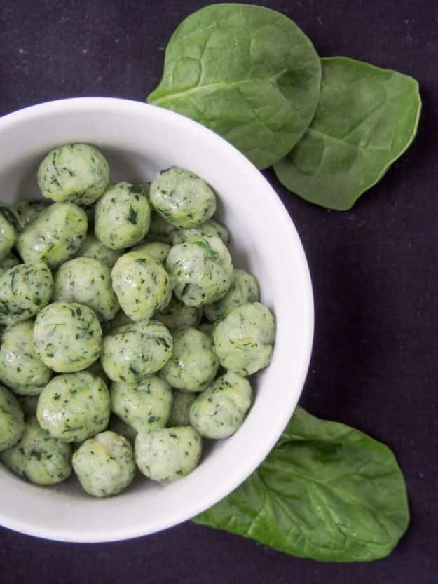 White bowl of pale green spinach gnocchi balls with whole fresh spinach leaves on a black background