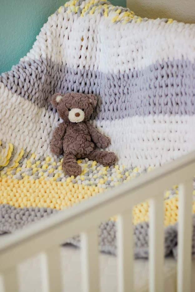 Knit baby blanket in white, gray and yellow draped over a crib rail with a small brown teddy bear sitting on it