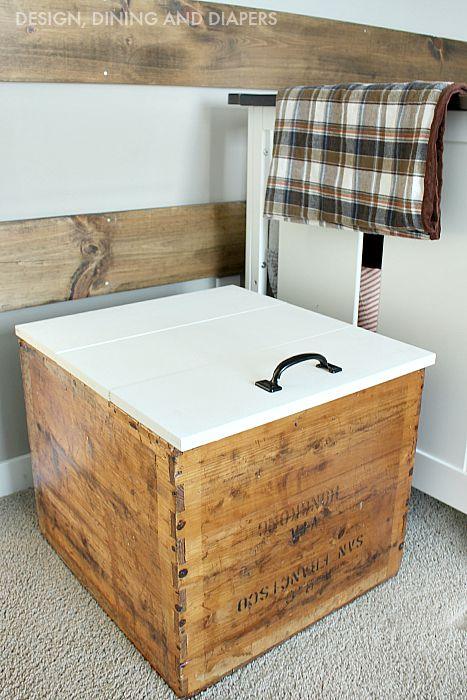 A vintage wooden shipping crate turned toy chest with a white painted plank lid and a black handle beside a plaid blanket