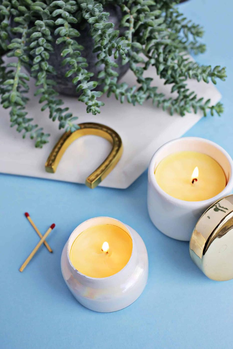 Two small white ceramic jar candles lit on a blue surface with a brass lid, matches and a trailing succulent