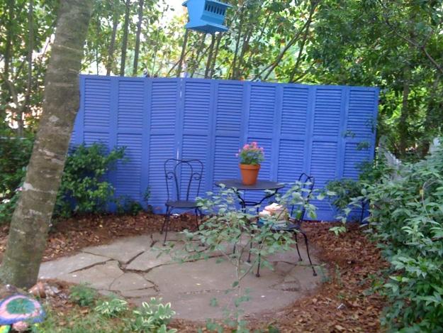Re-purposed shutter fence, blue painted louvred shutters standing as a fence around a patio corner with a bistro table