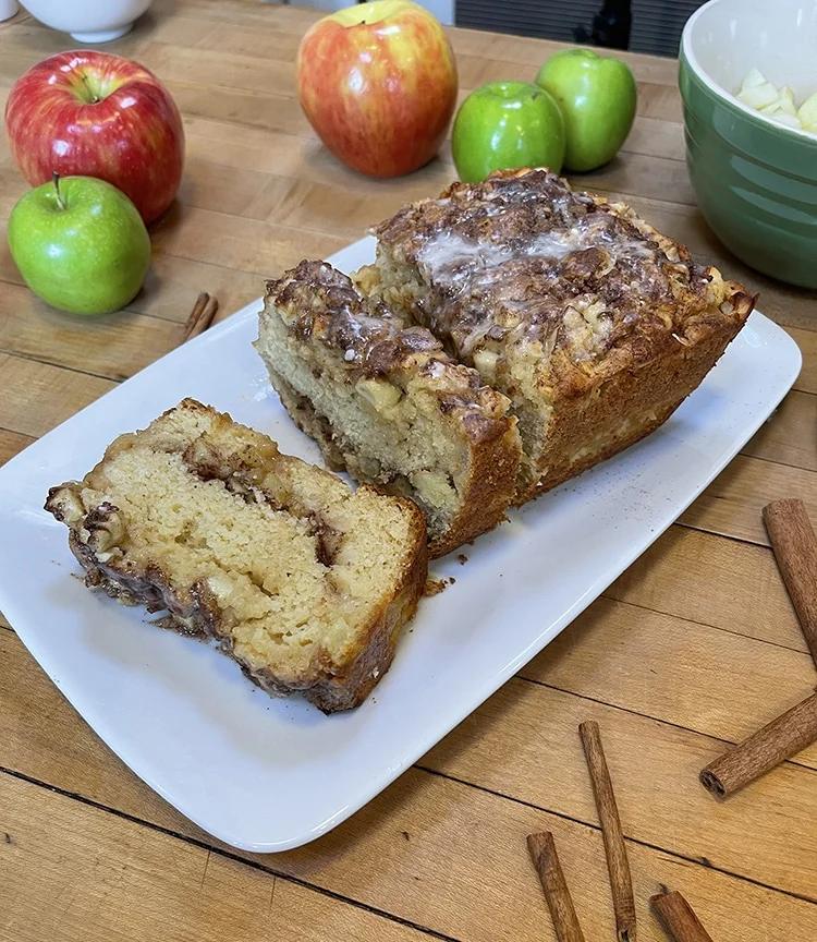Fall Baking - angled view of glazed cinnamon swirl apple bread slices on a white plate with red and green apples and a green bowl of chopped apple behind