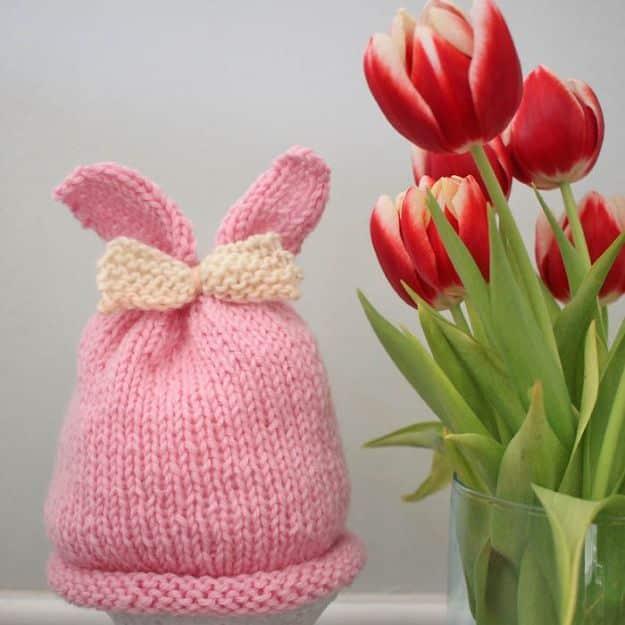 Pink baby bunny ear hat with a cream knitted bow at the base of the ears, standing beside a vase of red tulips