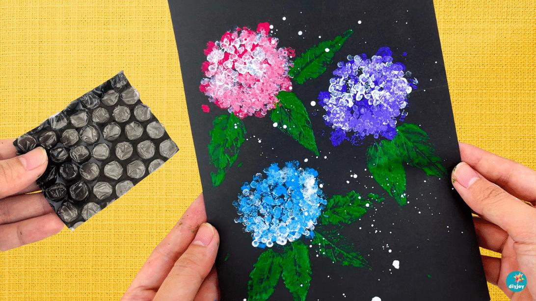 Hands pressing bubble wrap onto black paper to stamp pink, purple and blue hydrangea flowers with green leaves