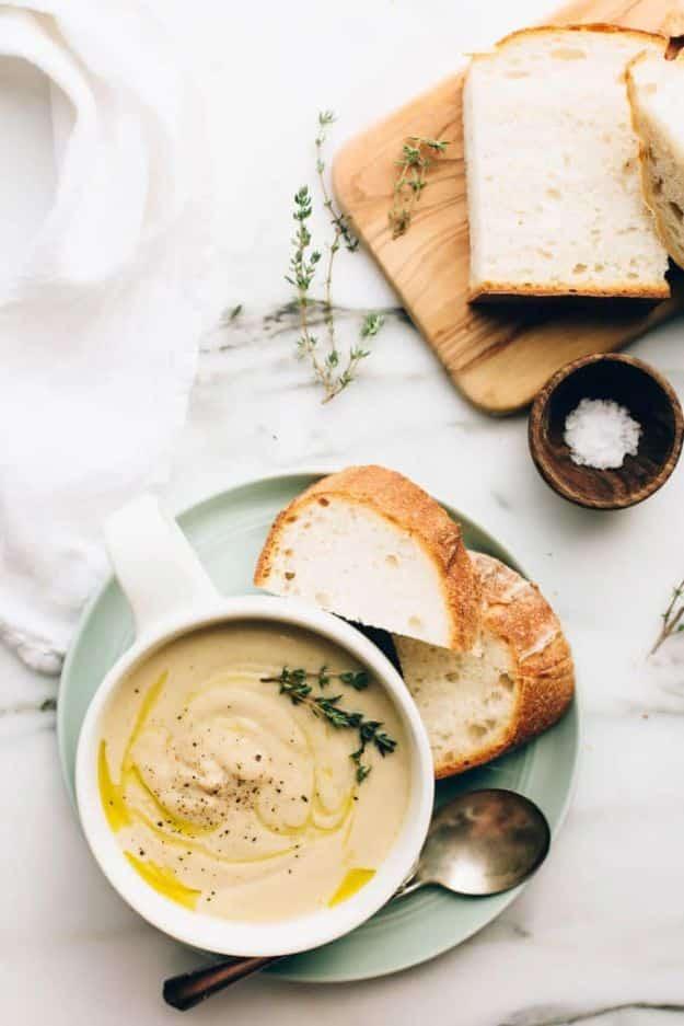 Creamy cauliflower soup in a white mug on a pale green plate with olive oil drizzle, thyme and sliced bread