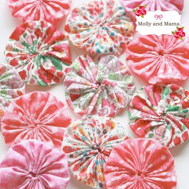Fourteen gathered fabric yo yos or Suffolk puffs in pink and coral floral prints laid out in rows on a white surface