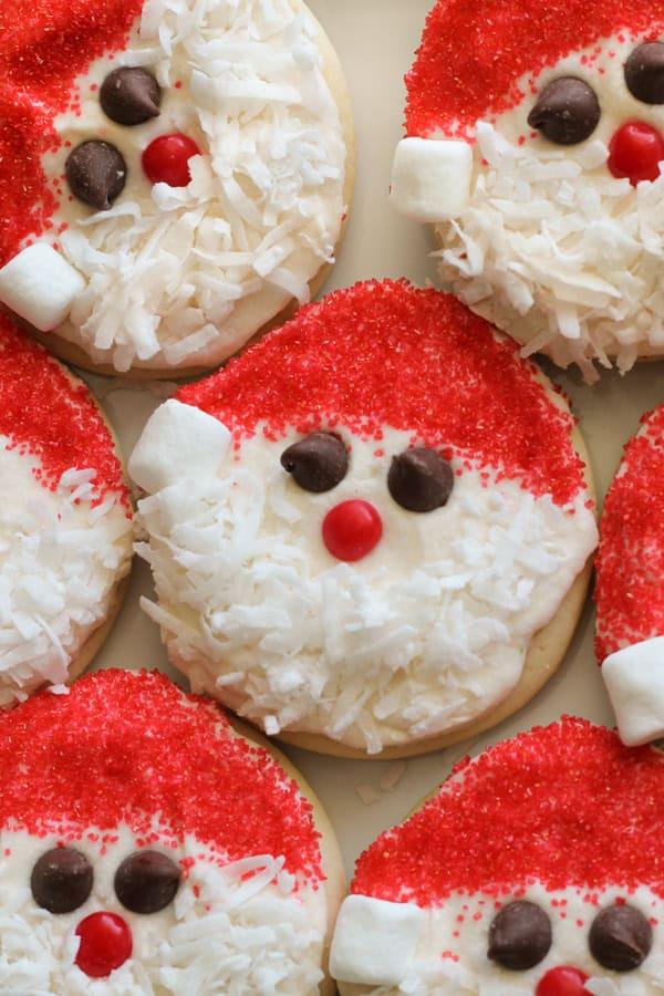 Round sugar cookies decorated as Santa faces with red sugar hats, marshmallow pom poms, chocolate chip eyes and coconut