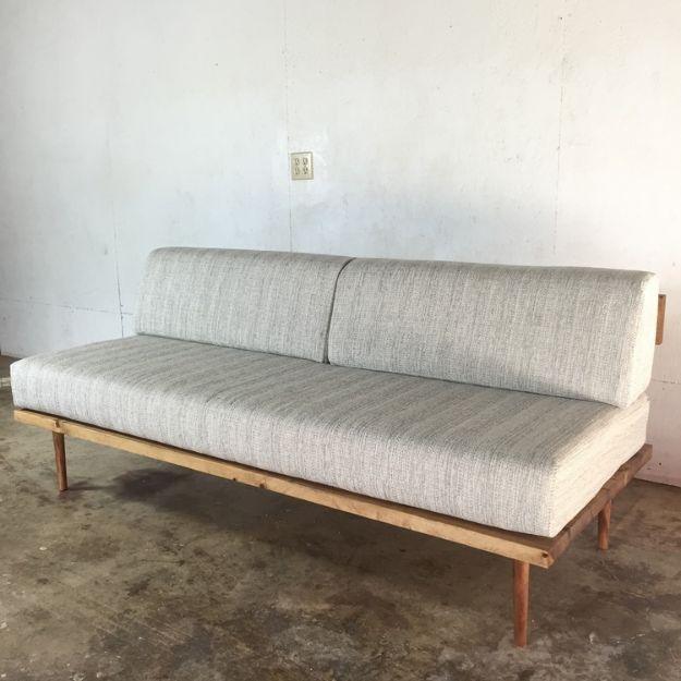 Low midcentury sofa with a pale walnut frame, tapered legs and two long grey woven cushions against a concrete wall