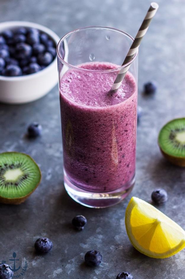 Deep purple blueberry lemon smoothie in a tall glass with a striped straw, kiwi, blueberries and a lemon wedge on slate