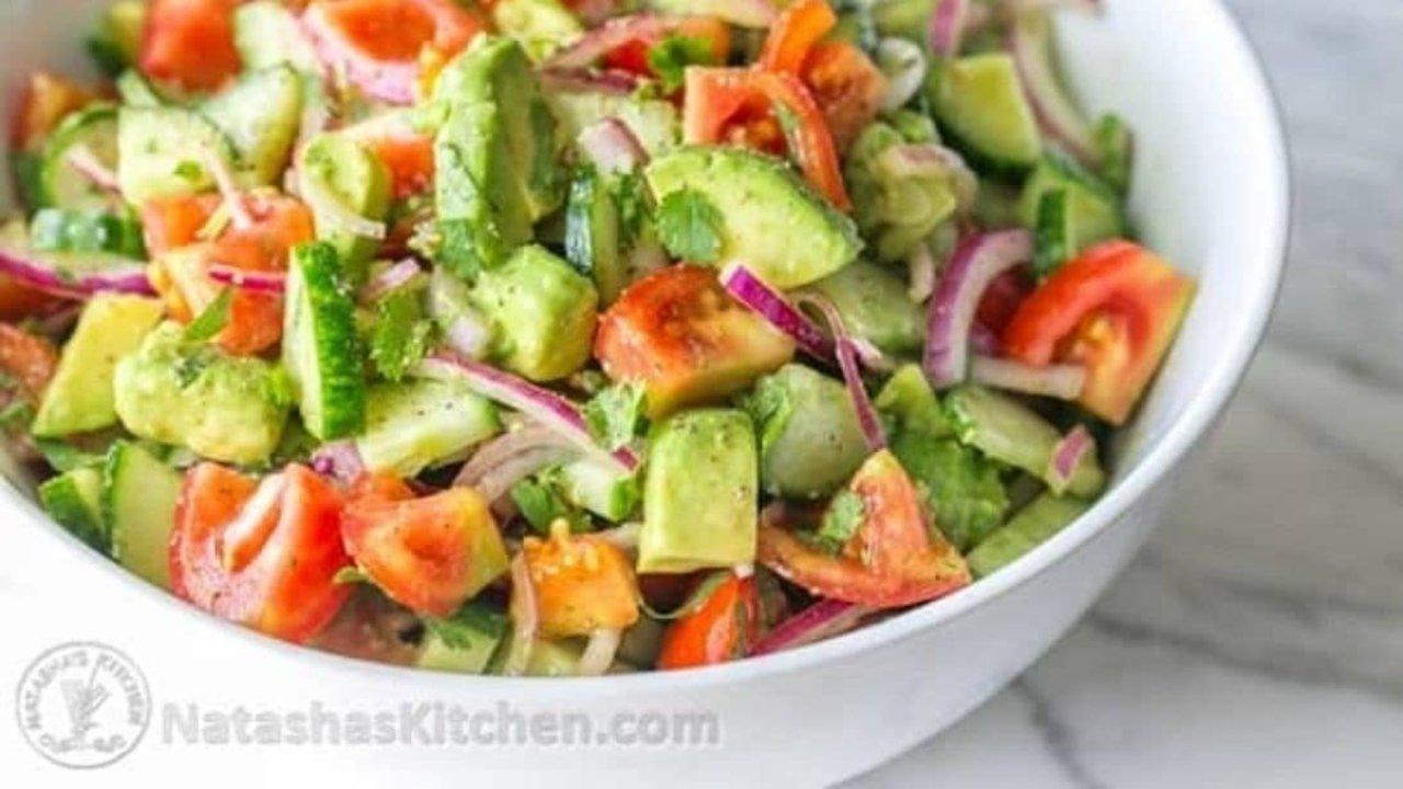 White bowl of cucumber tomato and avocado salad on marble, sliced red onion and chopped herbs, shot from above