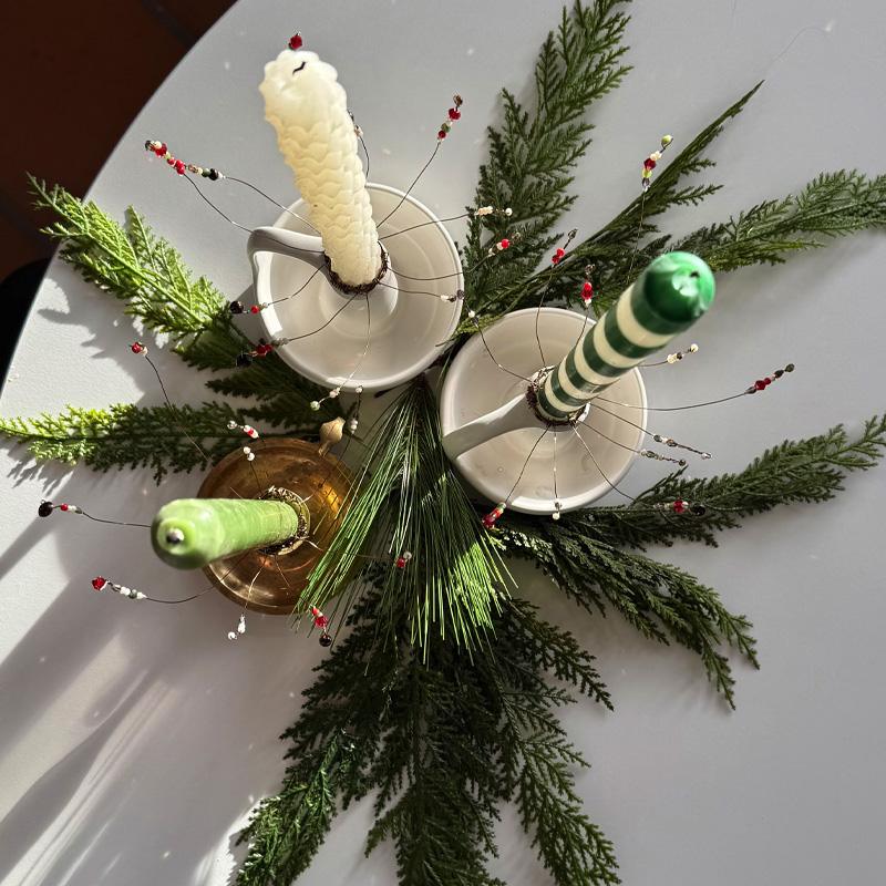 Taper candles in white holders on evergreen boughs, each wearing a braided wire cuff with red and silver bead sprays