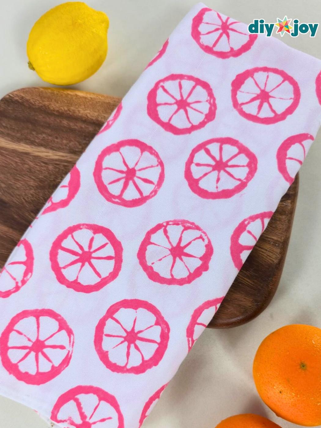 White flour sack tea towel stamped with pink citrus slice prints laid on a wood board beside a fresh lemon