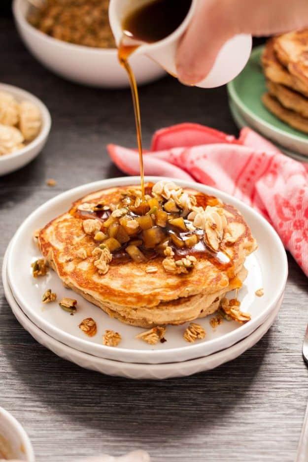 Apple cinnamon pancakes stacked on a plate with stewed apple pieces, granola and syrup being poured over