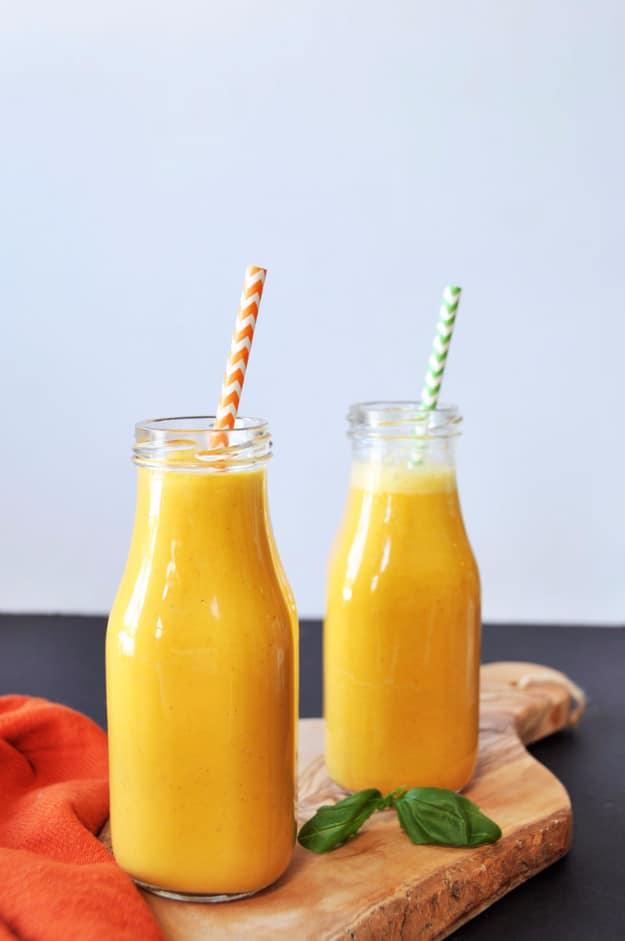 Two glass bottles of golden carrot turmeric ginger smoothie with a basil leaf on a wood board and orange napkin