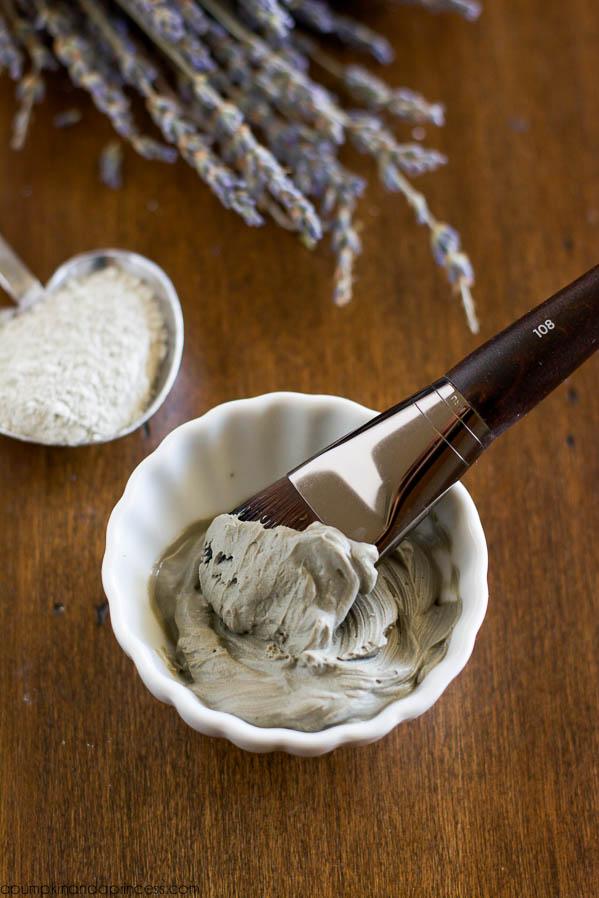 DIY clay face mask, a small white dish of grey clay paste with a flat makeup brush and dried lavender on dark wood