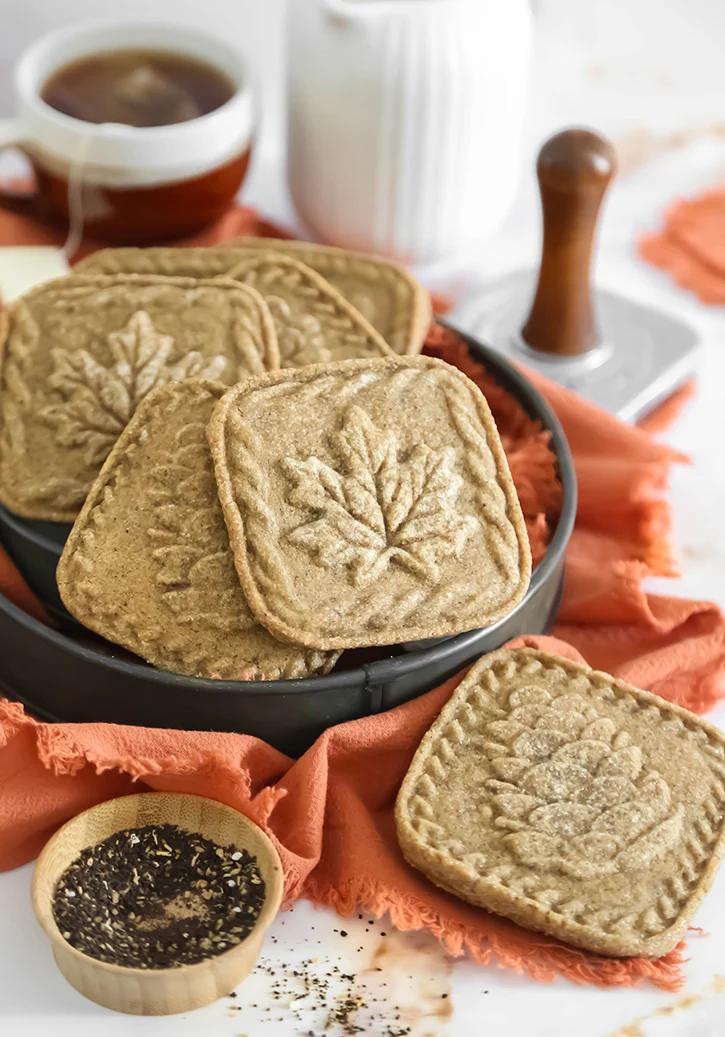 Square chai butter cookies stamped with maple leaf and pine cone designs in a tin on an orange napkin beside a cookie stamp