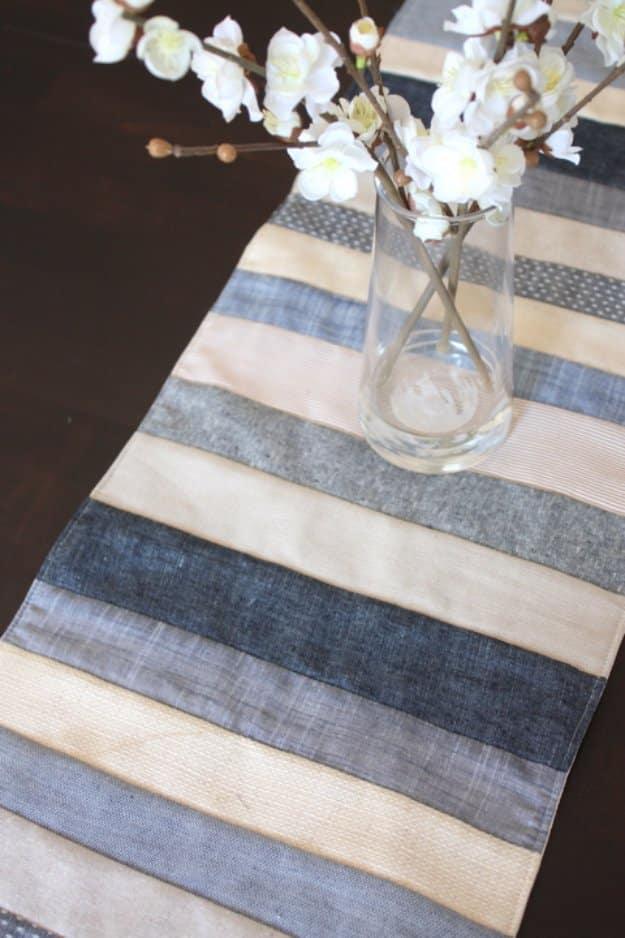 A pieced table runner of denim, chambray and linen strips in cream, oatmeal, gray and indigo down a bare wooden table.