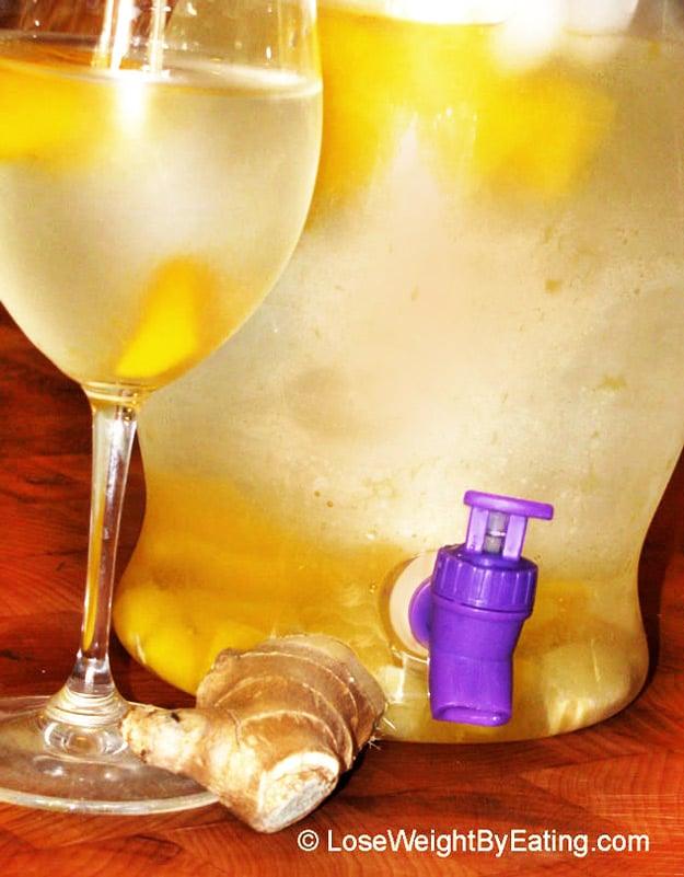 Mango ginger water in a glass drink dispenser with a spigot beside a wine glass and fresh ginger root