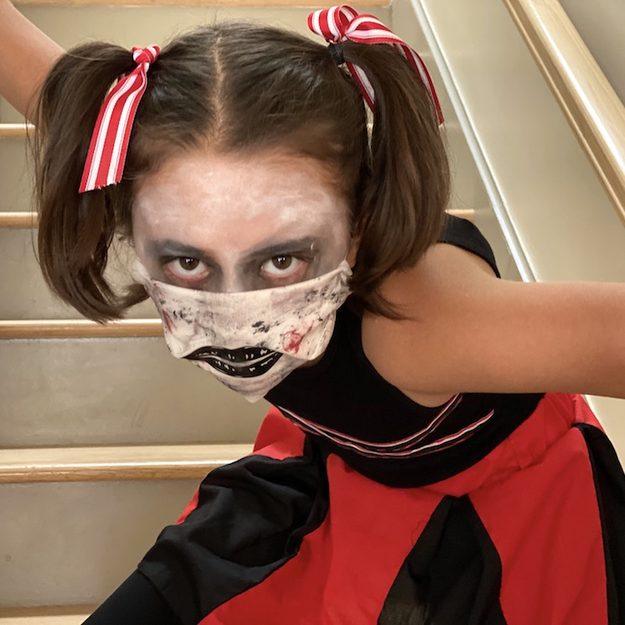 Zombie cheerleader Halloween makeup with pale skin, dark eyes and a painted fabric mask over the mouth