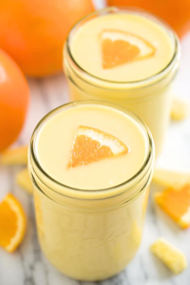Two mason jars of pale yellow pineapple orange banana smoothie topped with orange wedges beside oranges and pineapple