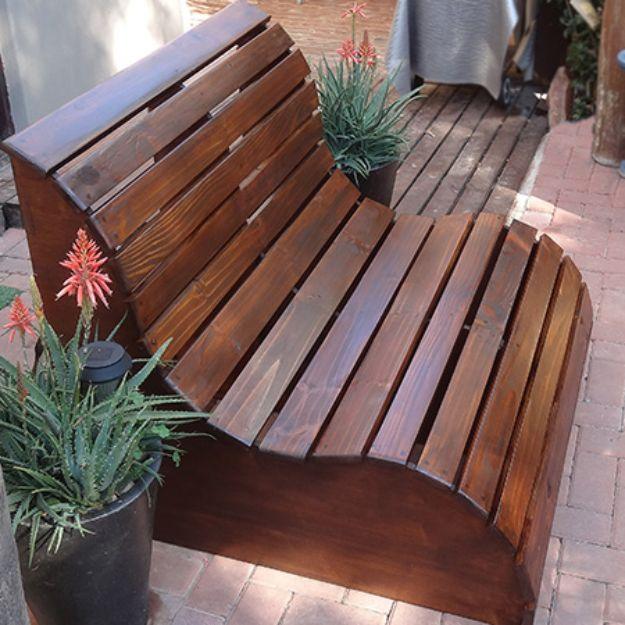Garden slat bench, a curved two seat lounger in dark stained wood slats on a brick patio beside potted aloes