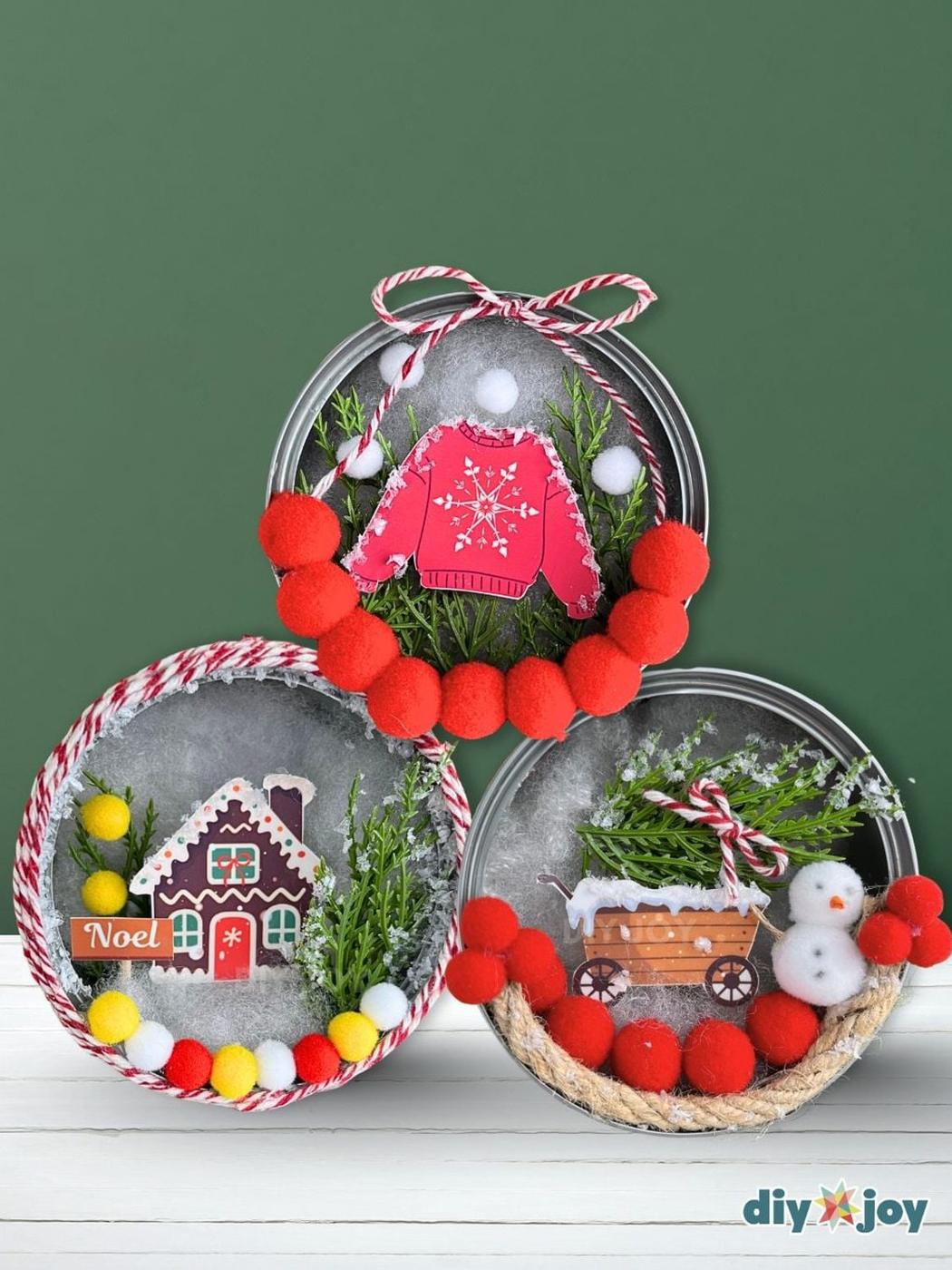 Three tuna can shadow box ornaments with a red sweater, a gingerbread house and a snowman scene, trimmed with pom poms