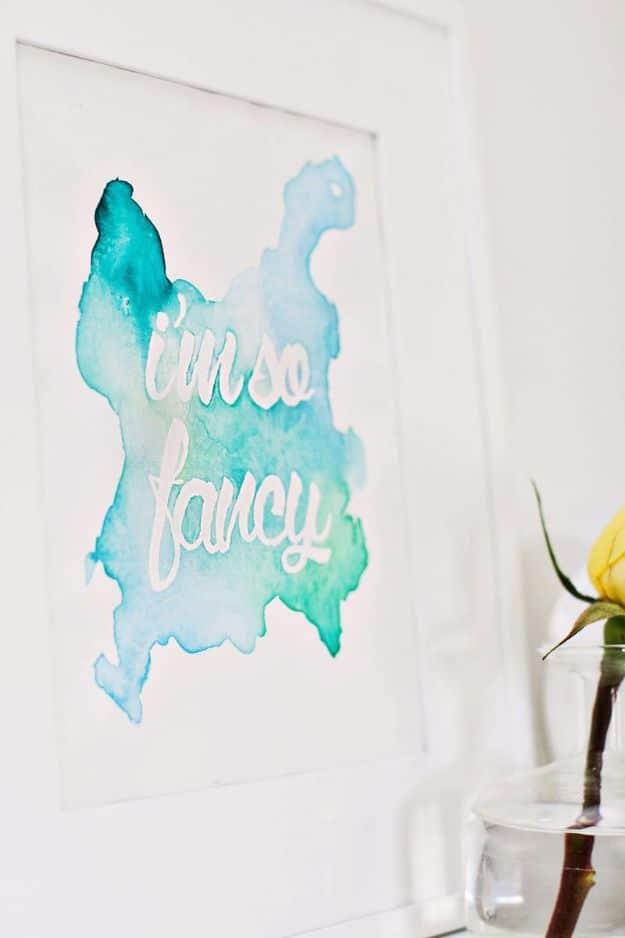 Framed watercolor wall art with a turquoise blot shape and white script lettering reversed out of it on a white wall