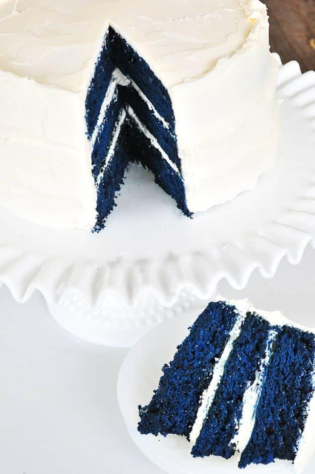 White frosted blue velvet cake on a pedestal stand with a cut slice showing three deep navy blue cake layers