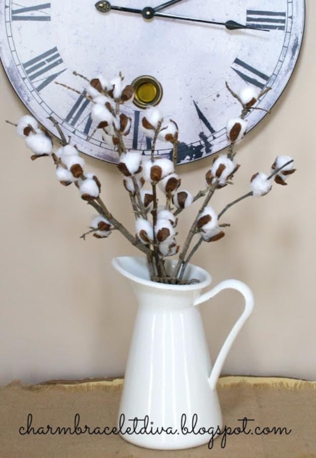 A bundle of faux cotton stems with white bolls on brown paper husks stands in a stoneware vase on a whitewashed shelf