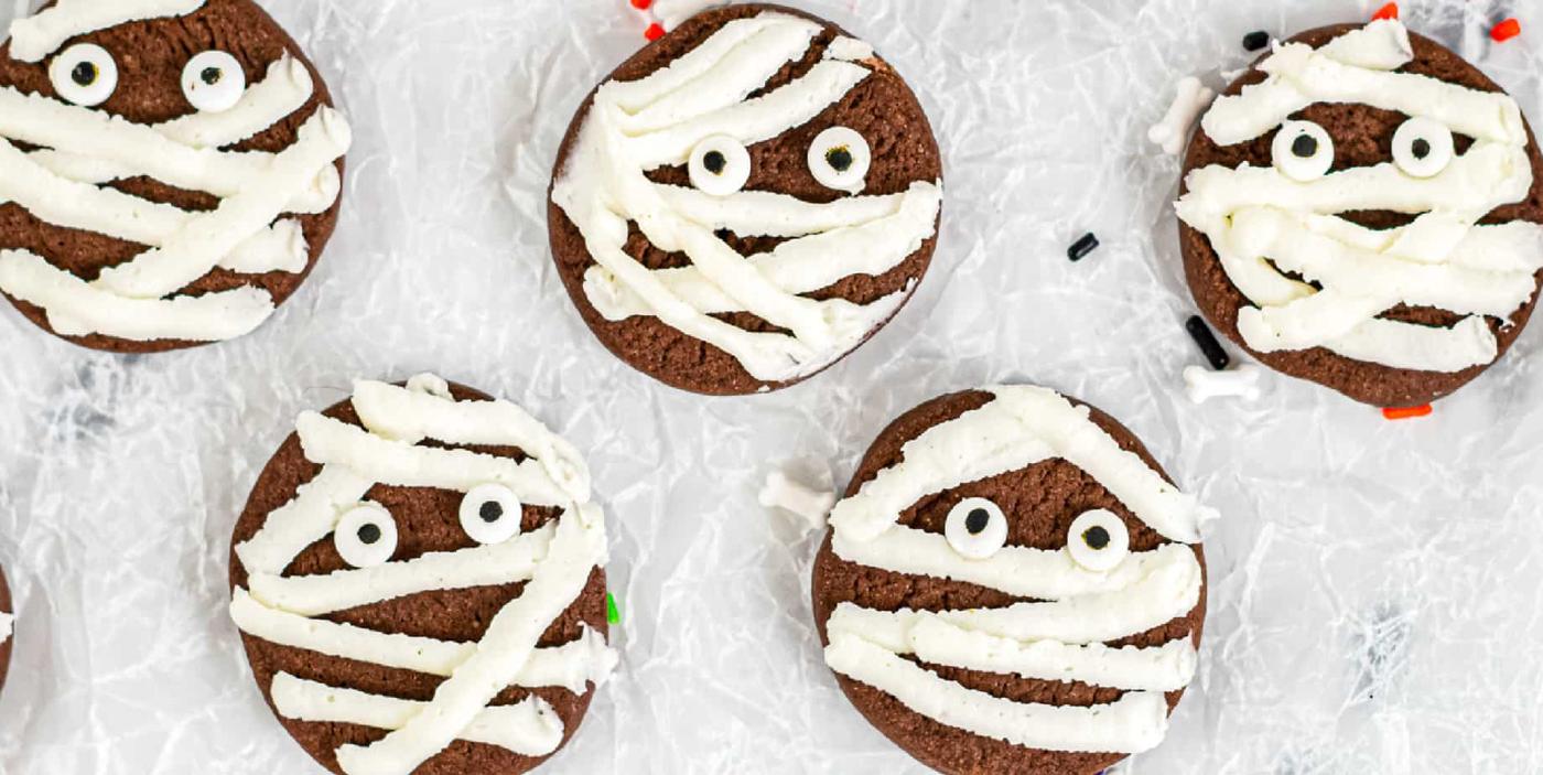 Small chocolate mummy cookies with white icing bandages and candy eyes arranged on a serving plate for Halloween