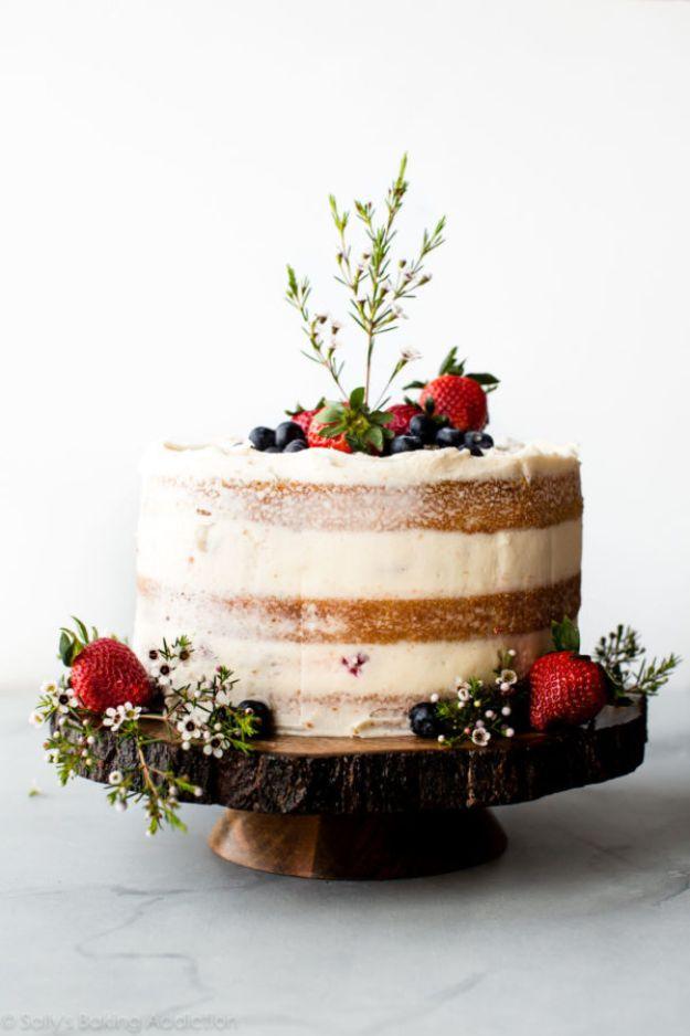 Vanilla naked cake with thin frosting showing the crumb, topped with fresh berries and herbs on a wood slab