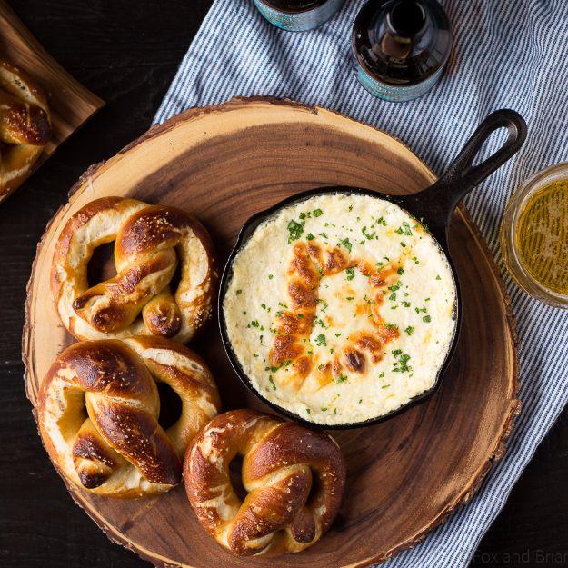 Soft beer pretzels on a wooden board beside a skillet of browned beer cheese dip topped with chives.