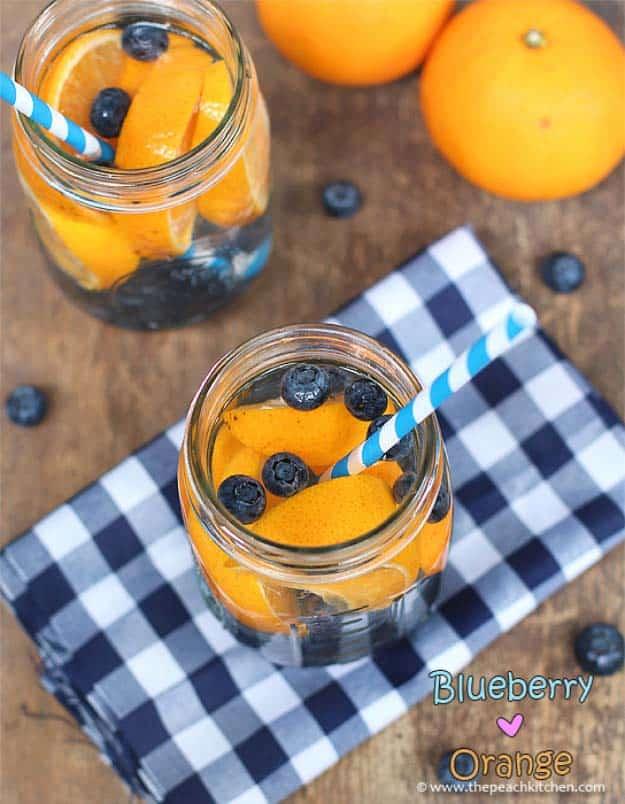 Blueberry and orange detox water in two jars with blue striped straws on a navy checked cloth with whole oranges