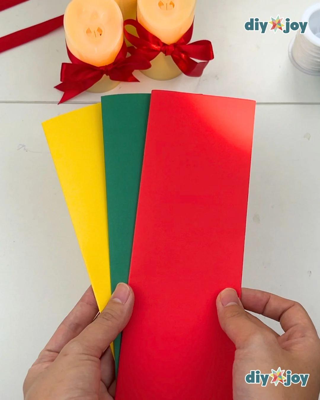 Paper Crafts - a hand holding up three flat sheets of yellow, green, and red cardstock fanned out in front of two lit candles
