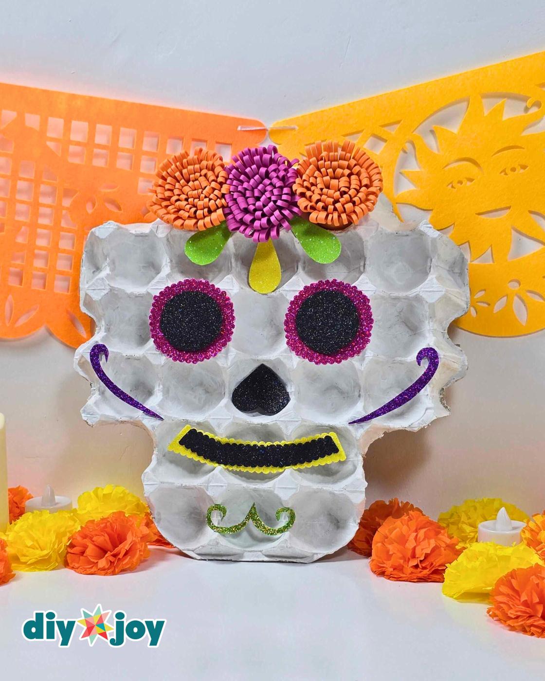 Recycled crafts decor - the egg tray skull standing in a white wall corner with orange and yellow die-cut papel picado, marigold paper flowers and a lit tea candle