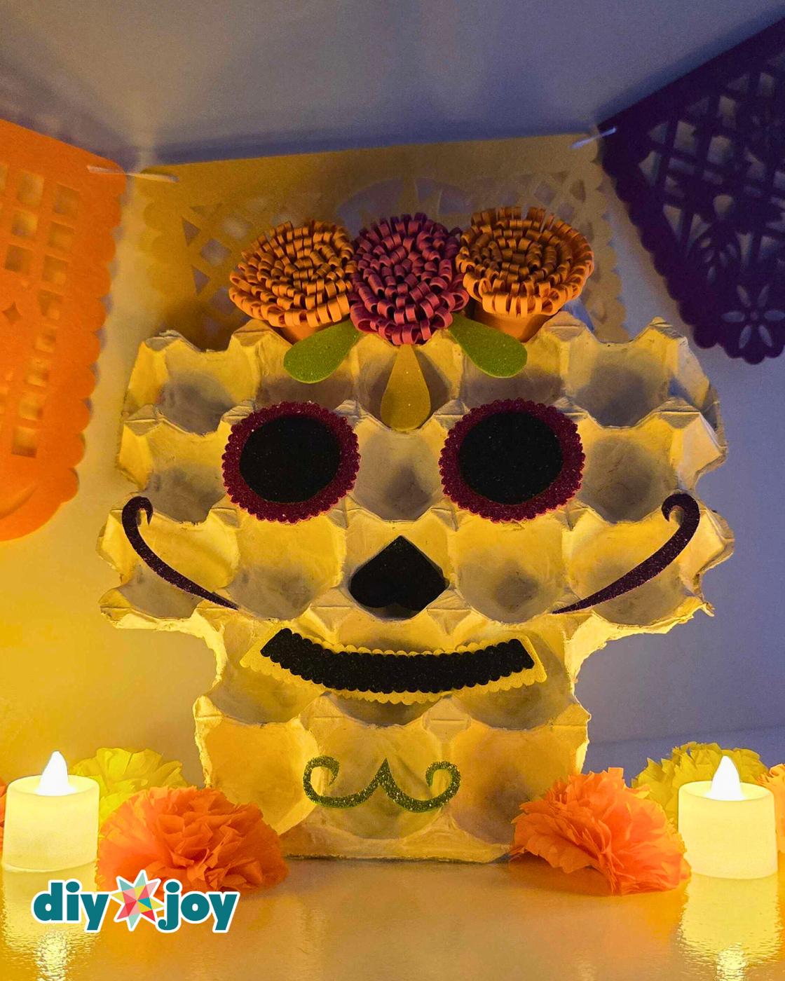 Day of the Dead crafts - the finished sugar skull propped upright on a shelf under warm lamp light with tea candles and paper marigolds at its base