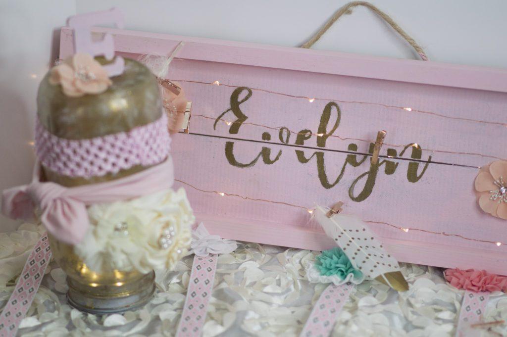 Pink painted wooden crate with the name Evelyn in gold, hanging ribbons, clothespin string lights and small bows