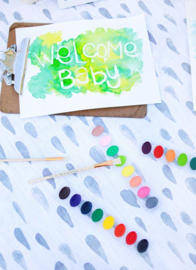 DIY baby shower decoration watercolor welcome baby sign on a clipboard beside two trays of paints and brushes