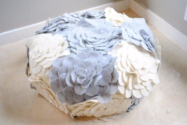 Round pouf covered in layered grey and cream felt flowers with hundreds of small petals, on a pale wood floor