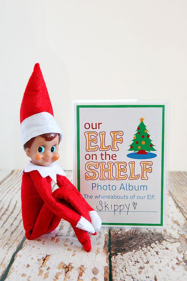 Elf on the Shelf doll leaning against a small green bordered photo album with a printed cover on white wood