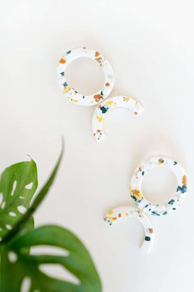 DIY terrazzo earrings in white polymer clay flecked with orange, teal and yellow, a hoop and crescent pair by a leaf