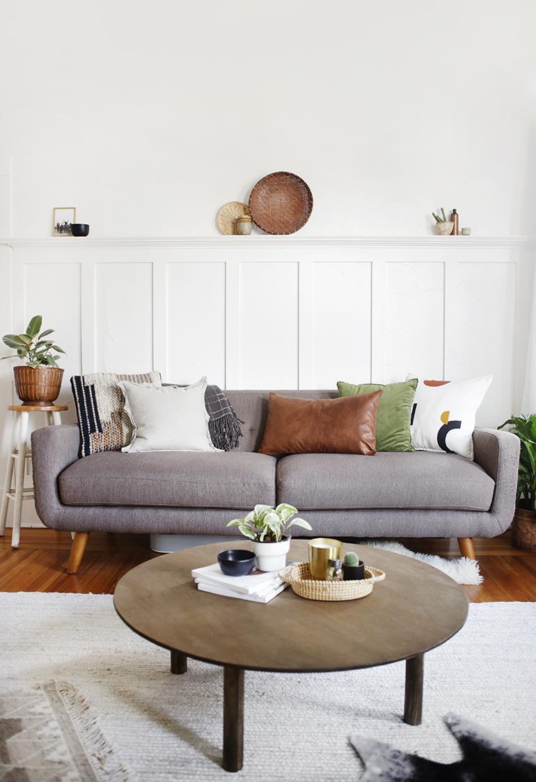 Gray fabric sofa with cushions in a neutral living room beside a round wood coffee table and a plant