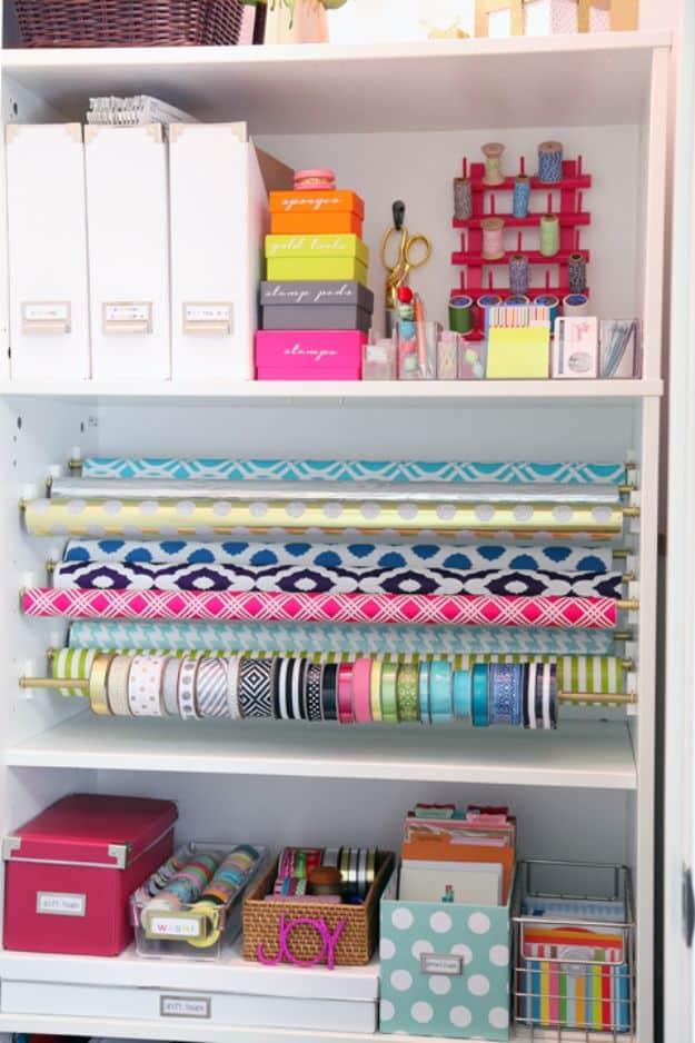 Bookcase gift wrap station with wrapping paper rolls on gold rods, ribbon spools below, labeled boxes and files above