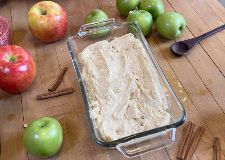 Cinnamon Apple Bread - pale unbaked batter smoothed into a clear glass loaf dish with a dark wooden spoon at the edge and apples and cinnamon sticks around it