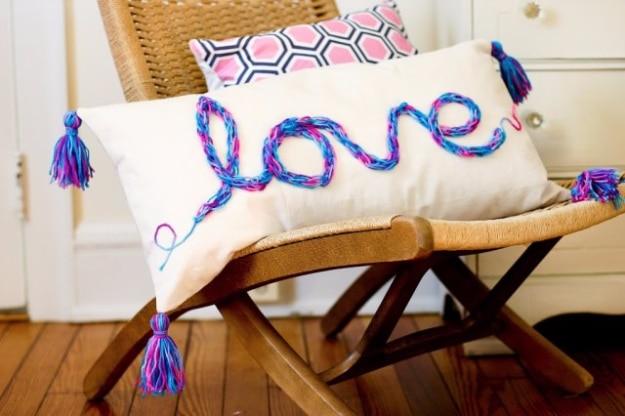 White canvas pillow with the word love in blue and pink finger knitting and four tassels, on a leather sling chair