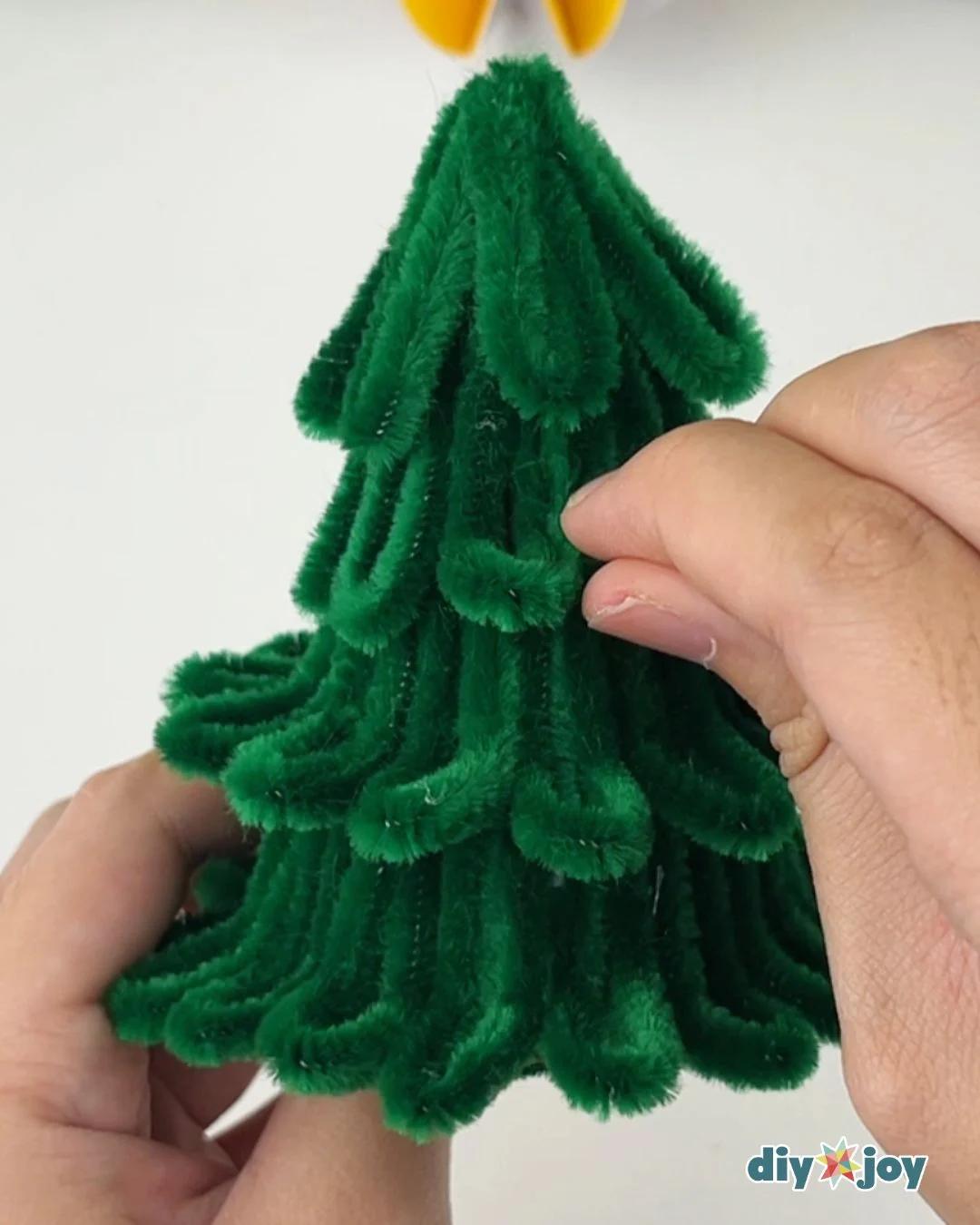 DIY Crafts - two hands curl the folded loop ends outward at the base of a fully covered green pipe cleaner cone on a white background