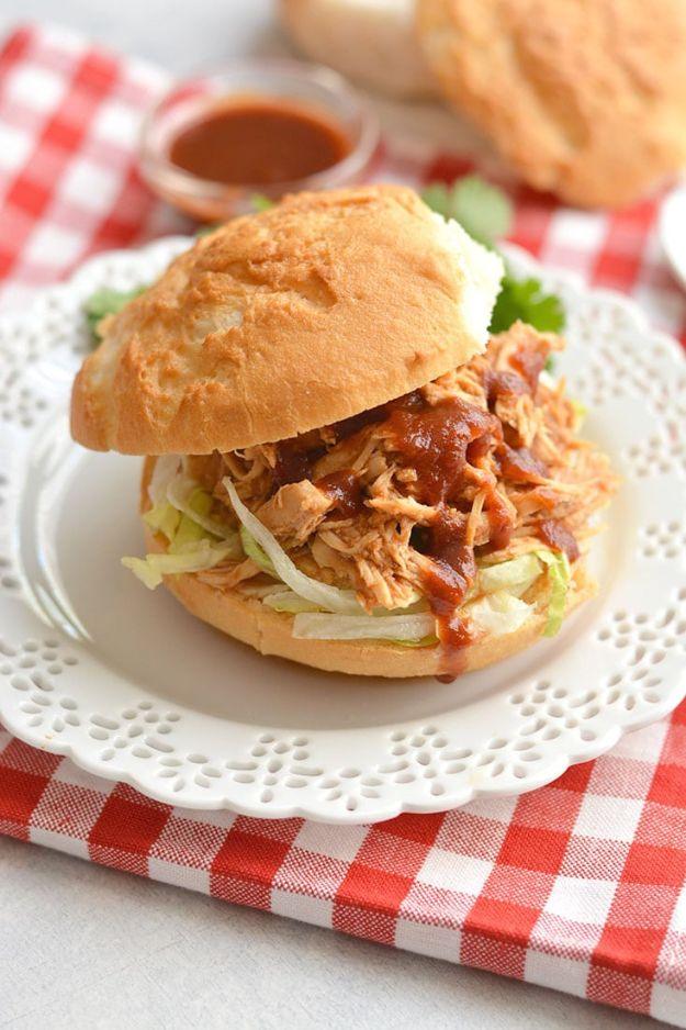 Healthy crockpot BBQ chicken piled on a bun as a sandwich, with a small bowl of barbecue sauce alongside