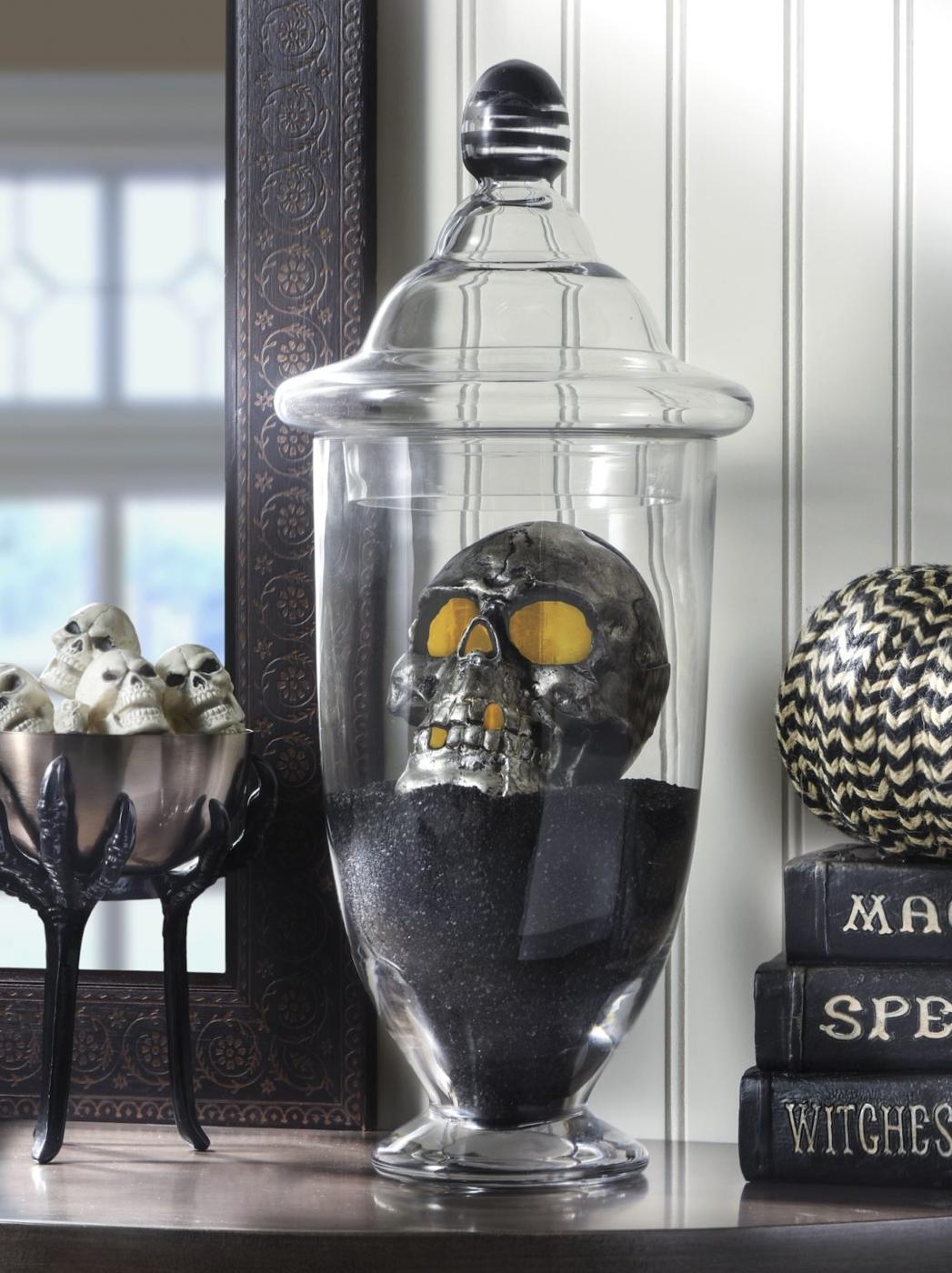 Black skull glowing inside a clear apothecary jar over black granules, with small skulls and spell books nearby