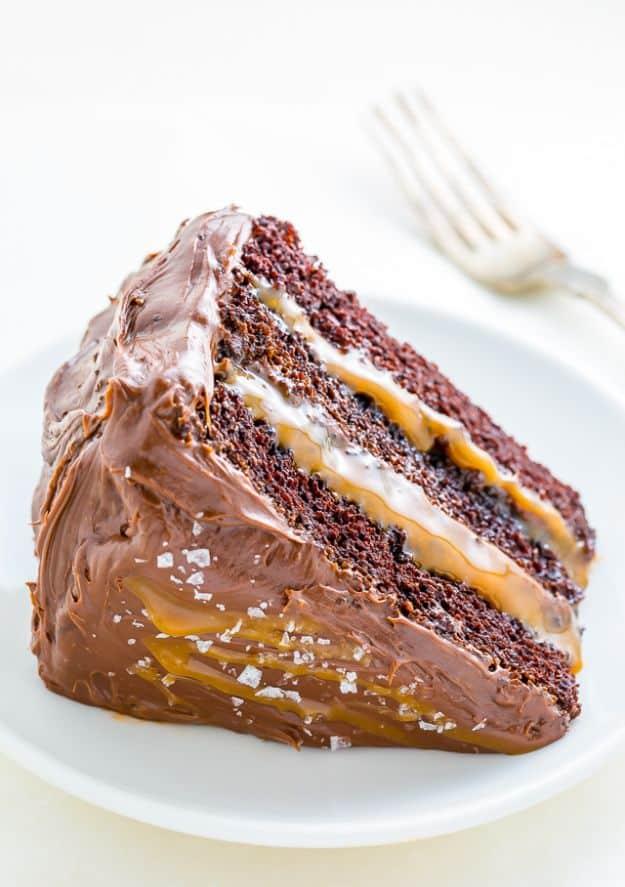 Salted caramel chocolate cake slice on a white plate with caramel running between layers and flaky salt